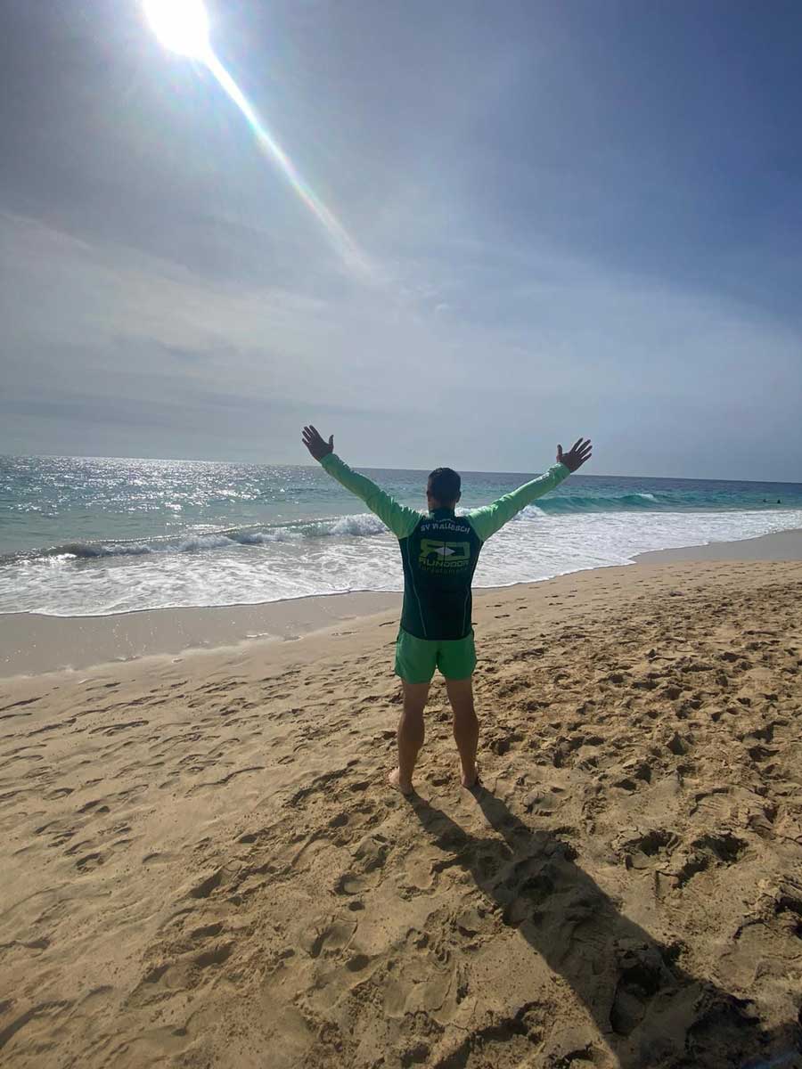 Trikot auf Tour - Mann mit ausgebreiteten Armen am Strand mit den Füßen im Sand, vor sich das Meer.