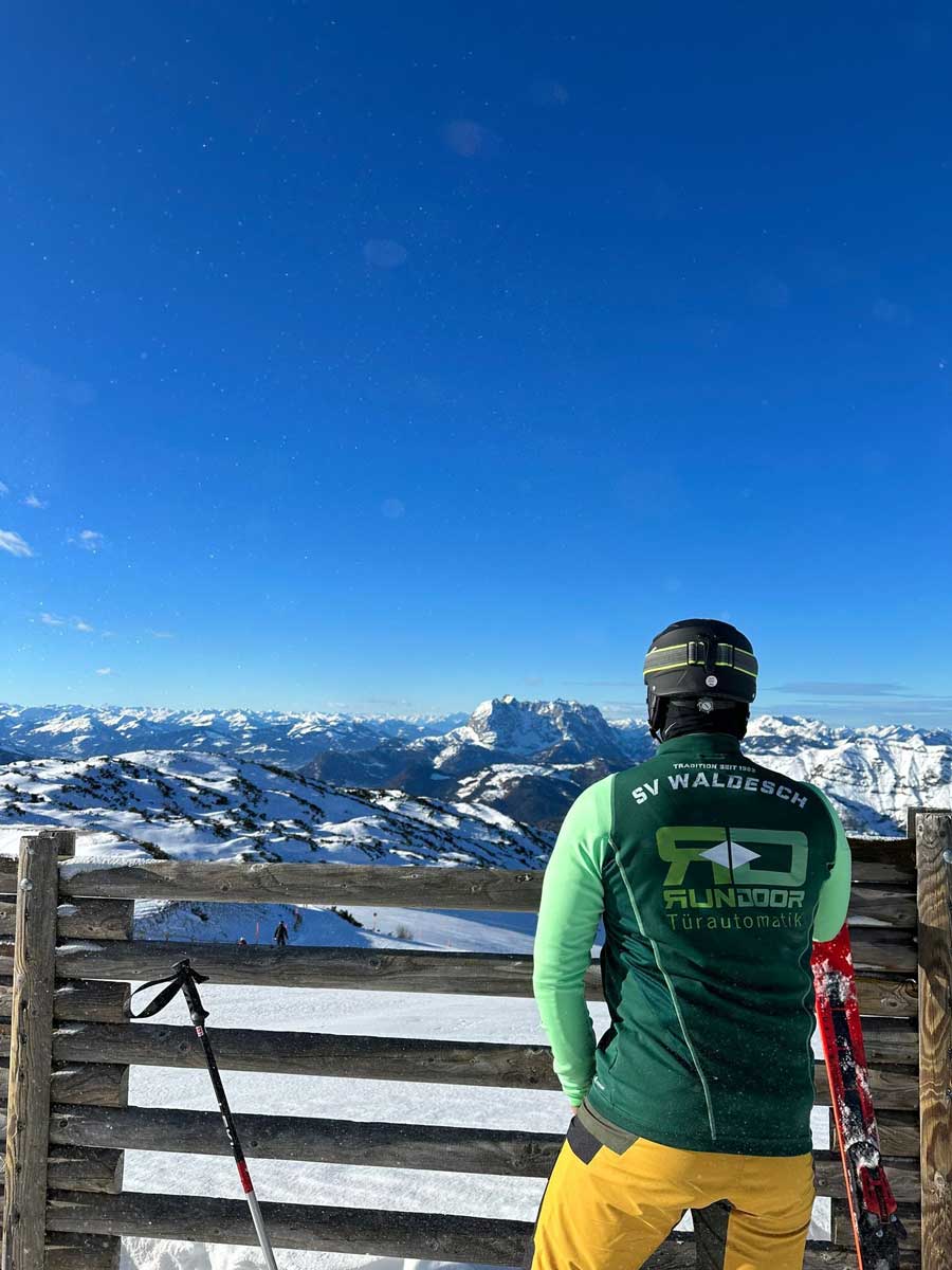 Trikot auf Tour - Junger Mann mit Skihelm hoch oben auf einem Gipfel mit Blick auf schneebedeckte Berglandschaft