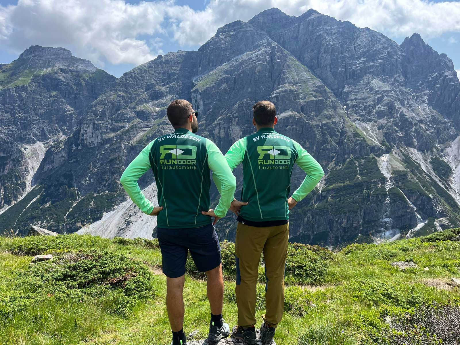 Trikot auf Tour - zwei Männer auf einer grasgrünen Wiese und Blick auf ein Felsmassiv in den Bergen